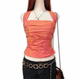 May include: A sleeveless, coral-coloured halter top with a ruched, iridescent fabric finish. The top is paired with dark blue denim jeans featuring a gold-toned chain belt with alternating square and circular links, and a gold pendant necklace.