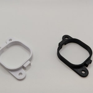 Könnte beinhalten: Zwei 3D-gedruckte Kunststoffhalterungen, eine weiße und eine schwarze, mit einer quadratischen Öffnung in der Mitte. Die Halterungen haben eine abgerundete Form und zwei kleine Löcher an der Unterseite.