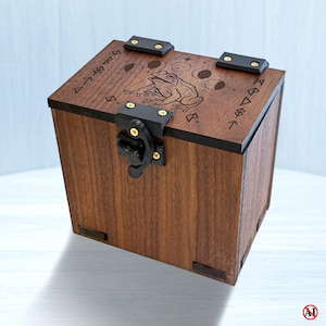 May include: A square wooden box with a dark brown finish, featuring a black metal latch and hinges. The lid is laser-engraved with a central frog illustration, a lunar cycle sequence, and geometric symbols along the edges of the top surface.