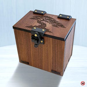 May include: A square wooden box with a dark brown finish featuring a laser-engraved bonsai tree design on the lid. The box includes black metal hinges and a black metal latch mechanism secured with small gold-colored screws on the front and top.