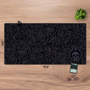 Può includere: Tappetino da gioco nero e grigio con motivo floreale, lungo 80 cm e largo 39,4 cm. Presenta un logo con teschio e fiamma nell'angolo. Sono visibili anche una piccola pianta e una carta di Magic: The Gathering.