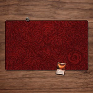 Può includere: Un tappetino da gioco rettangolare rosso intenso con un motivo floreale a spirale dettagliato. Un piccolo dado a più facce è appoggiato sul bordo del tappetino. Una carta collezionabile con un'immagine infuocata è posizionata sul tappetino.