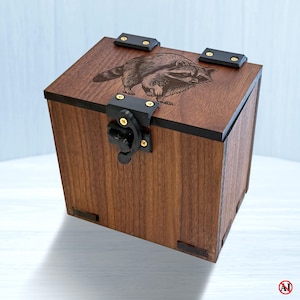 Peut inclure: Une boîte carrée en bois avec une finition brun foncé présentant un raton laveur gravé au laser sur le couvercle. La boîte comprend des charnières en métal noir et un loquet en métal noir fixé par de petites vis dorées sur les panneaux avant et supérieur.