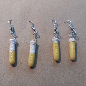 May include: Four pairs of novelty earrings. Each earring features a miniature test tube design, with a yellow body and a white top. The earrings are suspended from silver-colored fish hook ear wires.