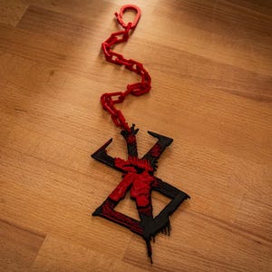 Op de afbeelding: Een rood en zwarte hanger met een ketting, met een gestileerd symbool. De hanger is een combinatie van rood en zwart, met een ketting van dezelfde rode kleur. Het ontwerp is geplaatst op een houten oppervlak.