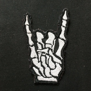 May include: White embroidered skeleton hand making the rock and roll hand gesture. The hand is on a black fabric background.