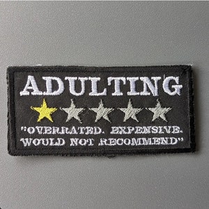 May include: Black embroidered patch with white text that reads "Adulting" and a one-star rating. The text below the rating reads "Overrated, Expensive. Would not recommend."