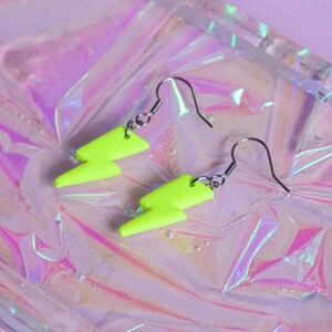 May include: A pair of neon green lightning bolt earrings with silver hooks. The earrings are on a iridescent pink background.