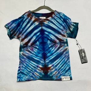 May include: A tie-dye t-shirt in blue, brown, and purple hues, hanging on a grey hanger. The shirt features a unique pattern. The tag reads "Manjacal Threadz".