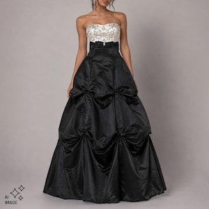 Op de afbeelding: Een strapless, vloerlange japon met een wit lijfje voorzien van gedetailleerd zilverkleurig borduurwerk en een zwarte rok met hoge taille. De rok is opgebouwd uit meerdere geplooide secties die een volumineus, gedrapeerd silhouet creëren.