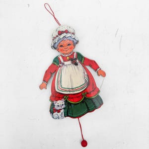 May include: A flat, hanging wooden ornament depicting a smiling character with grey hair wearing a white bonnet, a red dress with a white apron, and green trim. A small white kitten sits at the base of the dress. A red string is attached for hanging.