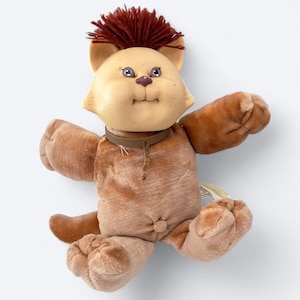 Peut inclure: Une peluche vintage en forme de chat avec un corps marron clair et un visage crème. Le chat a une crête brune, des yeux bleus et un collier marron. Le jouet est fait de tissu doux et a une expression douce.