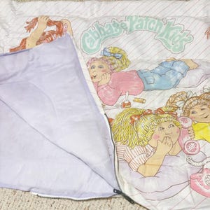 May include: A Cabbage Patch Kids sleeping bag featuring a white background with thin diagonal pink stripes. The graphic shows multiple dolls with blonde hair, blue jeans, and pink shirts. One doll holds a pink telephone. The interior is light purple.