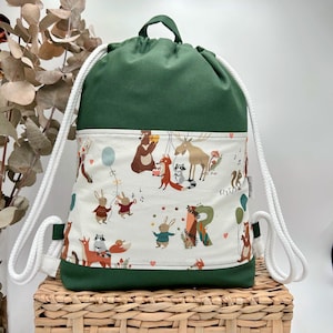 May include: A forest green drawstring backpack featuring a central white panel with a colorful print of woodland animals including bears, foxes, rabbits, and a moose, accented with white rope straps and a small fabric tag on the side.