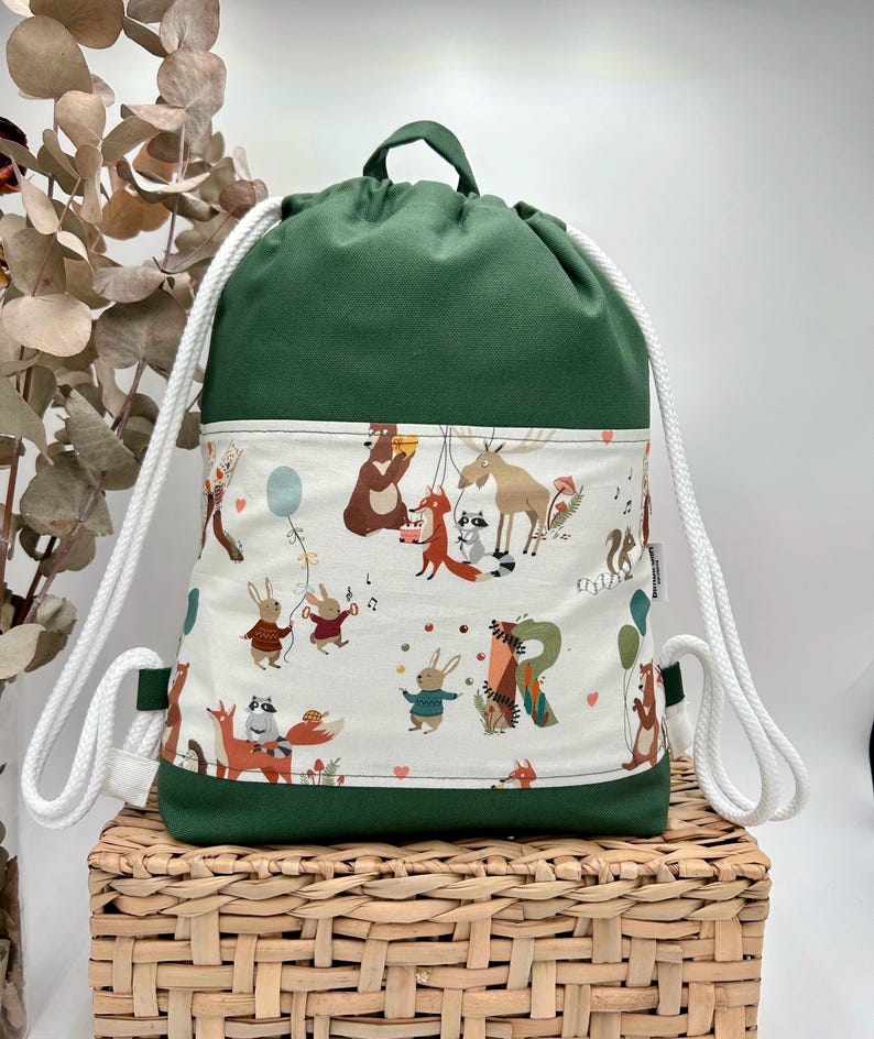May include: A forest green drawstring backpack featuring a central white panel with a colorful print of woodland animals including bears, foxes, rabbits, and a moose, accented with white rope straps and a small fabric tag on the side.