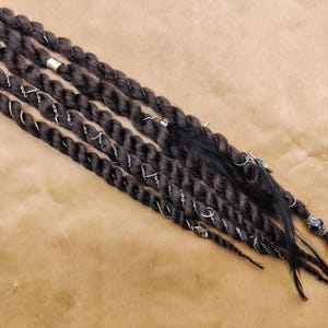 Puede incluir: Un conjunto de rastas sintéticas de color marrón oscuro con cuentas y abalorios plateados. Las rastas están dispuestas en fila sobre una superficie marrón.
