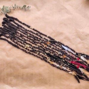 May include: A set of brown synthetic dreadlocks with gold beads, copper wire, and feathers. The dreadlocks are arranged in a row on a brown surface.