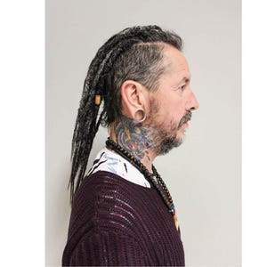 Op de afbeelding: Zijaanzicht van een persoon met grijze dreadlocks, een baard en een nek-tattoo. Ze draagt een donker bordeauxrode trui, een kralenketting en een wit overhemd met een bloemenpatroon.
