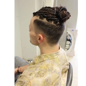 May include: A person with a bun hairstyle, featuring dark dreadlocks on top and a faded undercut. The person is wearing a light-coloured shirt with a floral pattern. The background includes a mirror and a white wall.