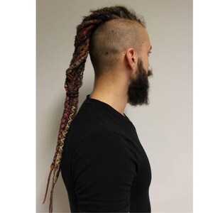 Puede incluir: Vista lateral de una persona con la cabeza rapada y un peinado de rastas largas y trenzadas. La rasta es una mezcla de colores marrón, rojo y rubio. La persona lleva una camiseta negra y tiene barba completa.