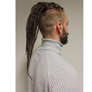 May include: A person with brown dreadlocks styled into a ponytail. The person is wearing a grey ribbed turtleneck jumper. The hair is shaved on the sides and back of the head, with a full beard.