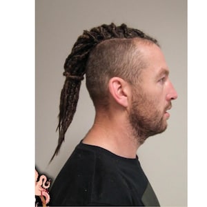 May include: A side profile of a man with a shaved head and a ponytail of dreadlocks. The dreadlocks are dark brown and tied at the base. The man is wearing a black t-shirt and has a beard.