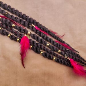 Puede incluir: Dreadlocks grises oscuros con cuentas doradas, plumas rosas y detalles dorados. Las rastas están dispuestas en diagonal sobre un fondo marrón. Las rastas están decoradas con anillos y dijes dorados.