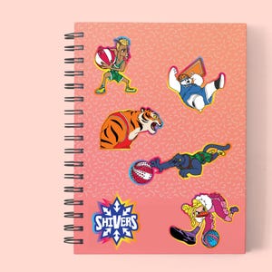 May include: Spiral-bound notebook with a pink cover featuring cartoon illustrations of athletes and animals playing sports. The notebook includes a basketball player, a tiger, a bear, a panther, and a woman. The word "SHIVERS" is printed on the cover.