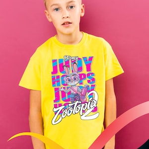 May include: A child wearing a yellow t-shirt with a colourful graphic of Judy Hopps from the Disney film Zootopia. The shirt displays the text "Judy Hopps" and "Zootopia 2". The background is a bright pink colour.