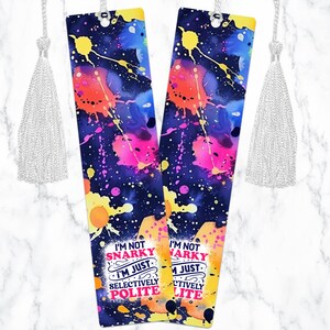May include: Two colorful bookmarks with a galaxy background and white paint splatters. The text on the bookmarks reads "I'm not snarky I'm just selectively polite".