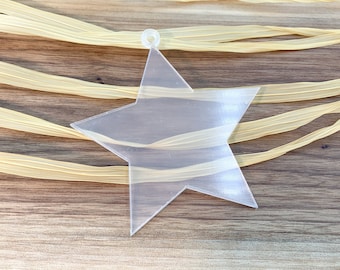 May include: A clear plastic star ornament with a hole at the top for hanging. The star is lying on a wooden surface with a light brown color.