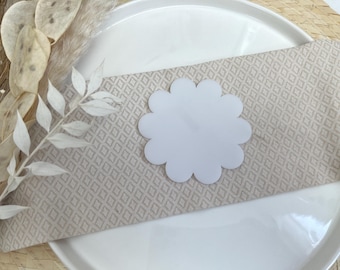 May include: A white, scalloped paper place card on a beige linen napkin with a diamond pattern. The napkin is on a white plate.