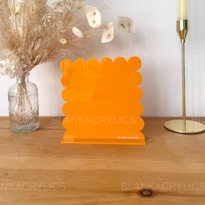 Peut inclure: Un présentoir en acrylique orange avec un bord festonné. Le présentoir a une petite base et est parfait pour présenter de petits objets. Le présentoir est fabriqué par Blank Acrylics.