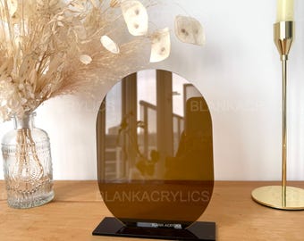May include: A brown oval-shaped acrylic mirror with a black base. The mirror is on a wooden surface with a bouquet of dried flowers and a candle holder in the background.