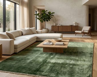 May include: A large rectangular area rug in a deep forest green hue with a subtle sheen and short fringe along the edges. The rug is positioned on a light-toned wooden floor in a room featuring a cream-colored sectional sofa and a wooden coffee table.