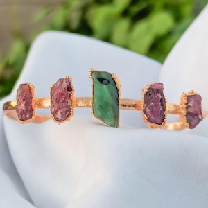 May include: A gold-coloured bracelet with five raw gemstone features. The gemstones are pink and green, each with a unique, irregular shape. The bracelet is displayed on a white surface with a blurred green background.