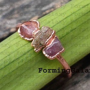 May include: A copper-colored ring featuring three raw, irregularly shaped gemstones in shades of pink and green. The ring is set against a green leaf and a wooden surface.