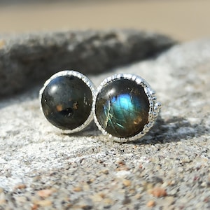 Peut inclure: Une paire de boucles d'oreilles à clous rondes avec des pierres précieuses de labradorite polies et sombres, serties dans des montures en métal argenté à bord perlé. Les pierres présentent des reflets bleus et verts irisés.