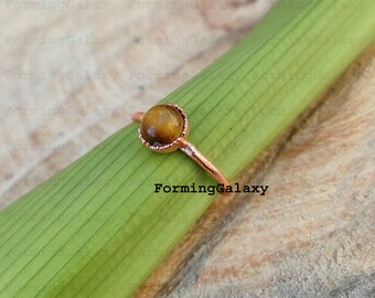 May include: A delicate ring with a round, golden-brown tiger's eye stone set in a copper-colored metal band. The ring is displayed on a green surface. The text "FormingGalaxy" is visible.