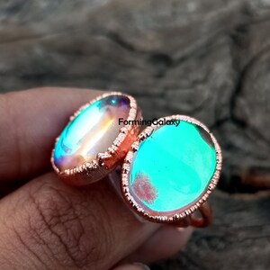 Puede incluir: Dos anillos con piedras ovaladas iridiscentes engastadas en metal color cobre. Una piedra tiene un tono azul-verdoso, la otra muestra un arcoíris de colores. Los anillos se muestran en primer plano, destacando los detalles.