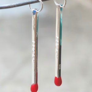 Può includere: Un paio di orecchini color argento a forma di fiammiferi. Ogni orecchino ha una punta rossa e la parola "LUCKY" incisa sul lato. Gli orecchini sono appesi a un filo d'argento.