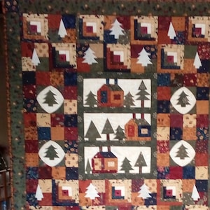 Könnte beinhalten: Eine handgefertigte Steppdecke mit einem rustikalen Thema "Hütte im Wald". Die Steppdecke zeigt Quadrate mit Blockhüttenblöcken, weißen Bäumen und Hüttenapplikationen in Braun-, Grün- und Rottönen. Der Rand ist dunkelgrün mit einem floralen Muster.