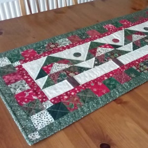 Peut inclure: Un chemin de table matelassé vert, rouge et blanc avec un motif d'arbre de Noël. Le chemin de table est fait de tissu patchwork et a un bord festonné.