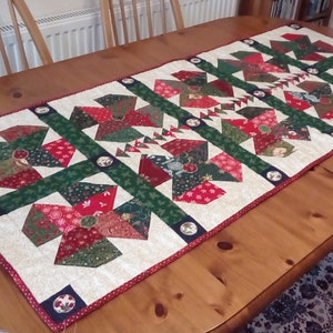 Op de afbeelding: Een witte tafelrunner met een kerstthema patchwork ontwerp. De runner heeft rode, groene en gouden stoffen met verschillende patronen. Hij is versierd met kleine, ronde, witte knopen.