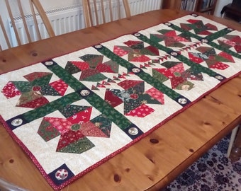 Peut inclure: Un chemin de table blanc avec un motif de patchwork sur le thème de Noël. Le chemin de table présente des tissus rouges, verts et dorés avec une variété de motifs. Il est décoré de petits boutons ronds blancs.