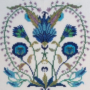 Può includere: Un ricamo a punto croce con un motivo a forma di cuore di fiori blu, verdi e viola con dettagli intricati.