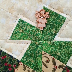 K&ouml;nnte beinhalten: Ein gr&uuml;ner und goldener Stoff-Patchworkblock mit einem h&ouml;lzernen Engels-Anh&auml;nger in der Mitte. Der Patchworkblock besteht aus vier Diamantenformen, jede mit einem anderen Muster. Der Engels-Anh&auml;nger ist ein schlichtes Design mit einem Heiligenschein und Fl&uuml;geln.