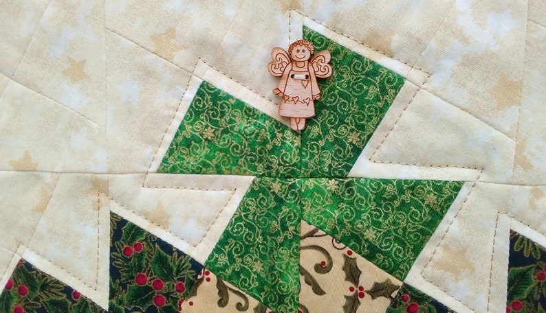 K&ouml;nnte beinhalten: Ein gr&uuml;ner und goldener Stoff-Patchworkblock mit einem h&ouml;lzernen Engels-Anh&auml;nger in der Mitte. Der Patchworkblock besteht aus vier Diamantenformen, jede mit einem anderen Muster. Der Engels-Anh&auml;nger ist ein schlichtes Design mit einem Heiligenschein und Fl&uuml;geln.