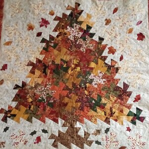 K&ouml;nnte beinhalten: Eine Steppdecke mit Patchwork-Muster, die einen gro&szlig;en Baum aus verschiedenen Braunt&ouml;nen, Orange- und Gr&uuml;nt&ouml;nen zeigt. Der Baum ist von kleineren Bl&auml;ttern und Blumen in Wei&szlig;-, Gelb- und Braunt&ouml;nen umgeben.