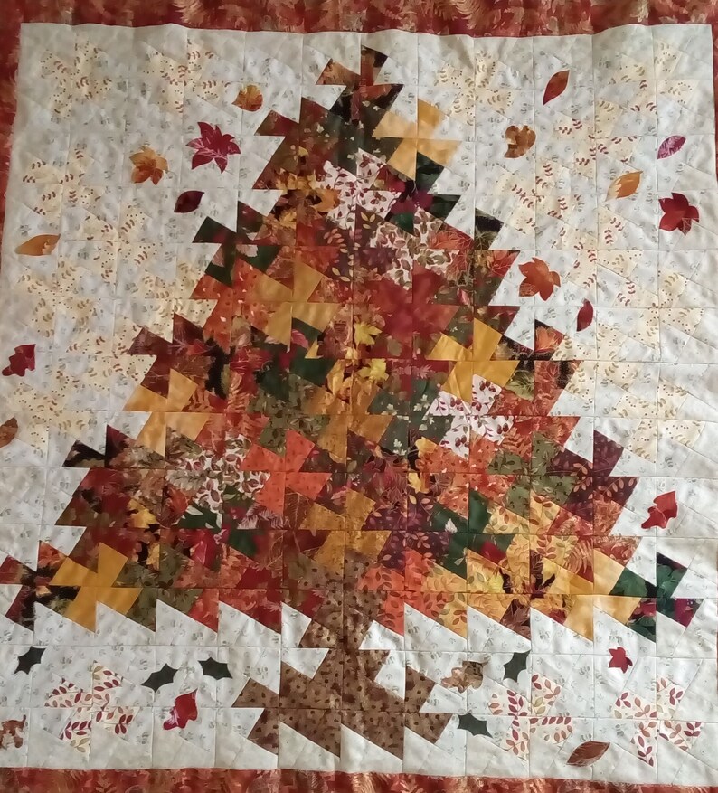 K&ouml;nnte beinhalten: Eine Steppdecke mit Patchwork-Muster, die einen gro&szlig;en Baum aus verschiedenen Braunt&ouml;nen, Orange- und Gr&uuml;nt&ouml;nen zeigt. Der Baum ist von kleineren Bl&auml;ttern und Blumen in Wei&szlig;-, Gelb- und Braunt&ouml;nen umgeben.